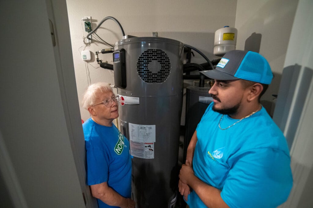 A plumbing technician and a homeowner stand together inside a utility closet inspecting a tall gray storage tank water heater.