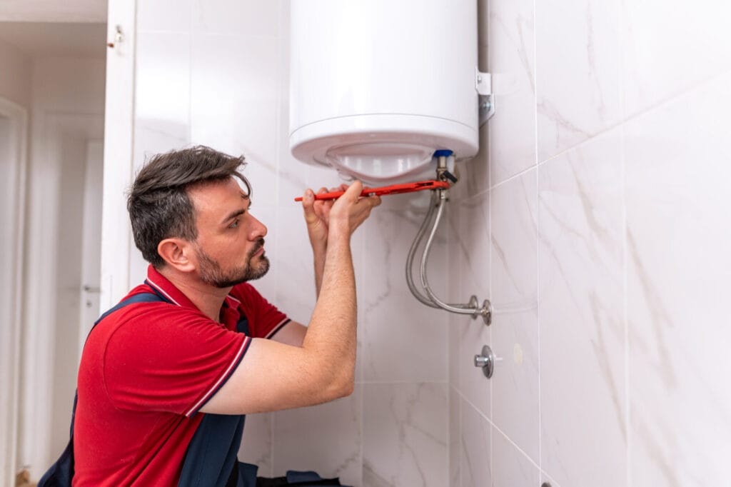 A plumber in a red shirt uses a wrench to adjust a white tank while performing Water Heater Troubleshooting.