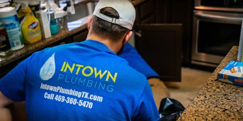 A professional plumber from Intown Plumbing wearing a blue company shirt works under a kitchen sink to address a clog.