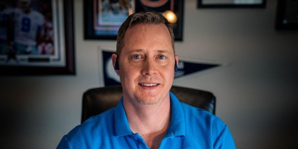 A professional headshot of a man wearing a blue polo shirt and a headset, positioned in an office setting with sports memorabilia in the background.