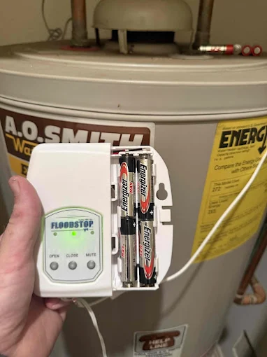 A hand holds a white, battery-powered Floodstop device in front of a large water heater to demonstrate a smart water leak detector setup in Rockwall TX.