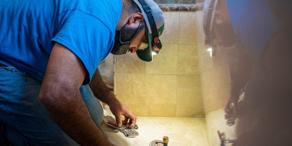An Intown Plumbing technician wearing a headlamp and mask kneels on a tiled floor to perform a plumbing repair.