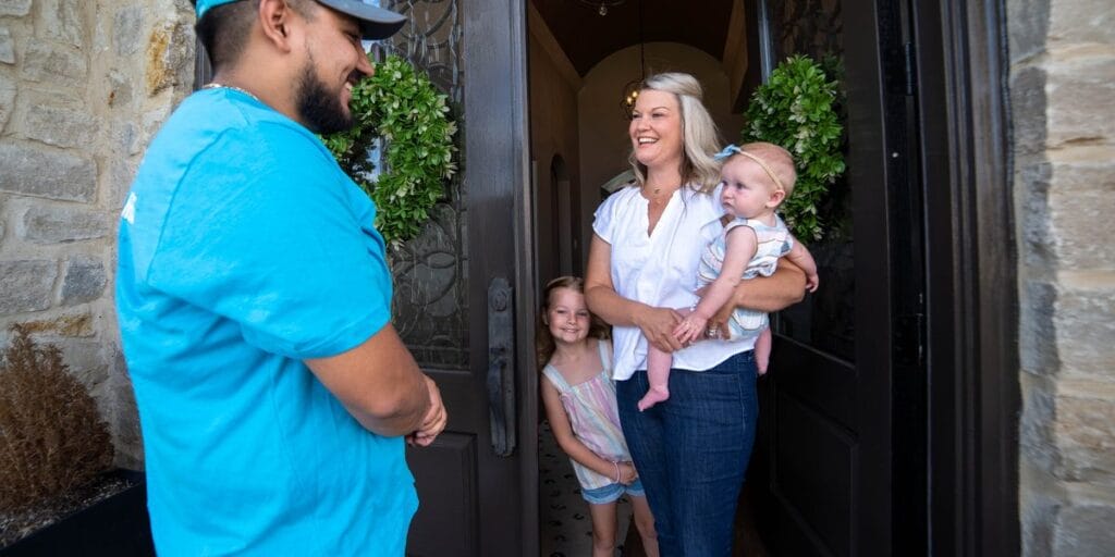 A technician in a teal polo shirt stands at a homeowner's open front door, greeted by a smiling woman holding an infant while a young girl peeks out beside her.
