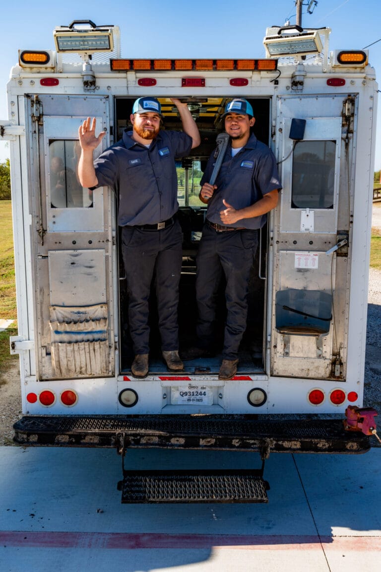 Two professional plumbers in matching gray uniforms and blue caps stand inside the back of a utility truck, with one waving and the other holding a large pipe wrench.