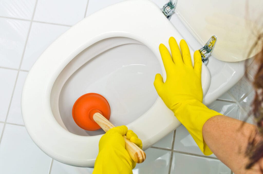 Person wearing yellow gloves using a plunger to unclog a toilet, demonstrating a method to clear blockages.