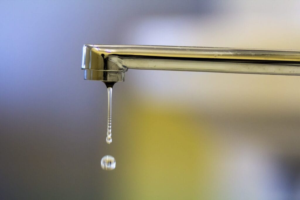 A close-up of a chrome faucet with a single drop of water hanging from the spout, with a second drop falling below it.