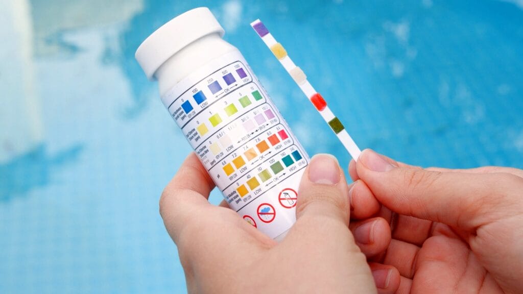 A hand holding a water test strip and a bottle with a color chart, used to check water quality by detecting different contaminants.
