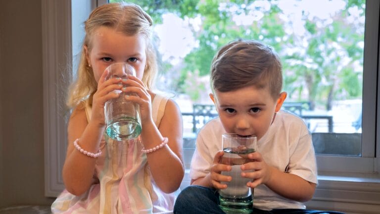 Two children drinking clean water from glasses, highlighting the importance of safe and tested drinking water.