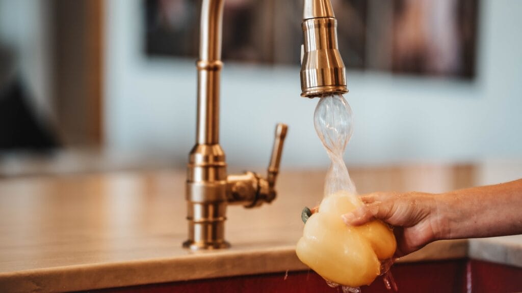 A person washing a yellow bell pepper under a modern kitchen faucet