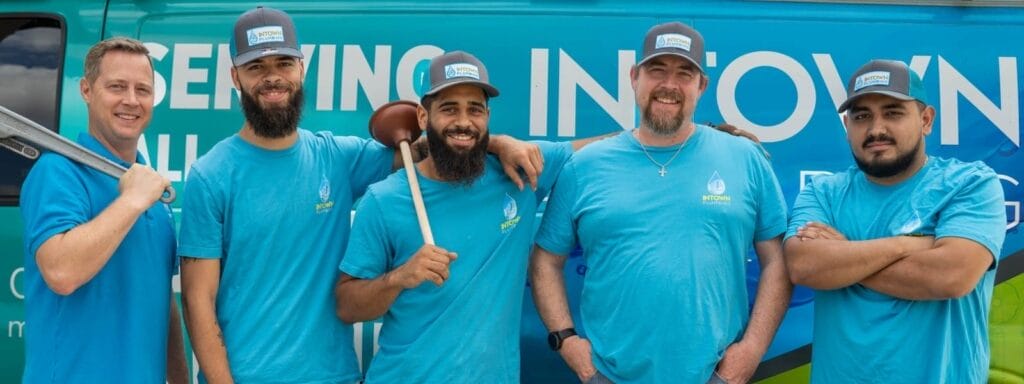 Five plumbers in matching blue uniforms stand smiling in front of a company-branded plumbing van. One plumber holds a plunger, while another holds a large pipe wrench, showcasing their tools and friendly team dynamic
