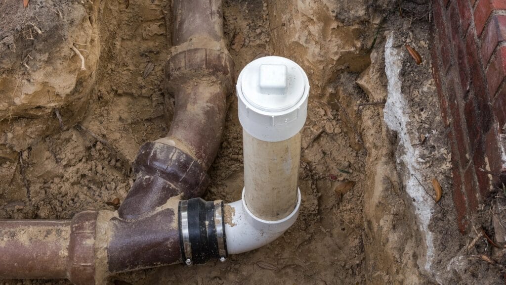 A close-up view of an outdoor sewer cleanout pipe with a white plastic cap connected to larger brown sewer pipes, partially buried in soil near a brick wall.