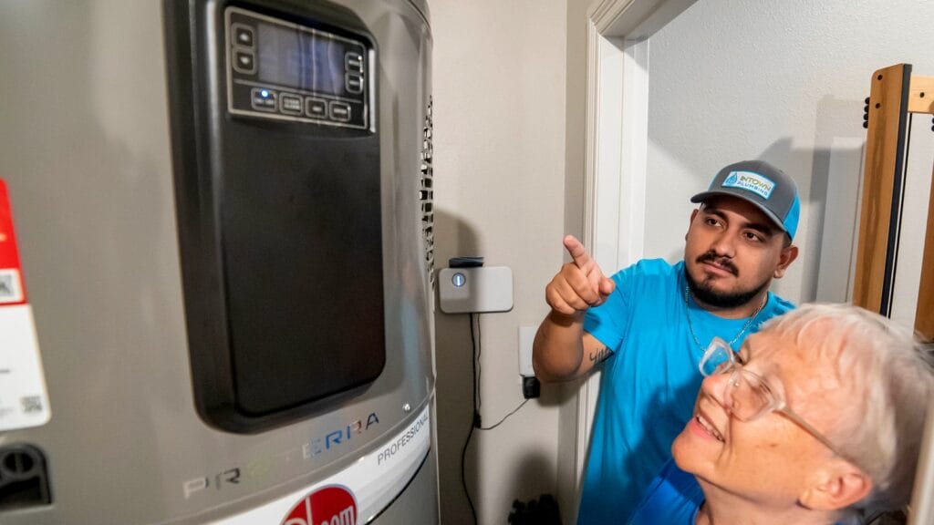 A plumber in a blue shirt and cap explains a water heater's settings to an elderly customer, pointing at the control panel.