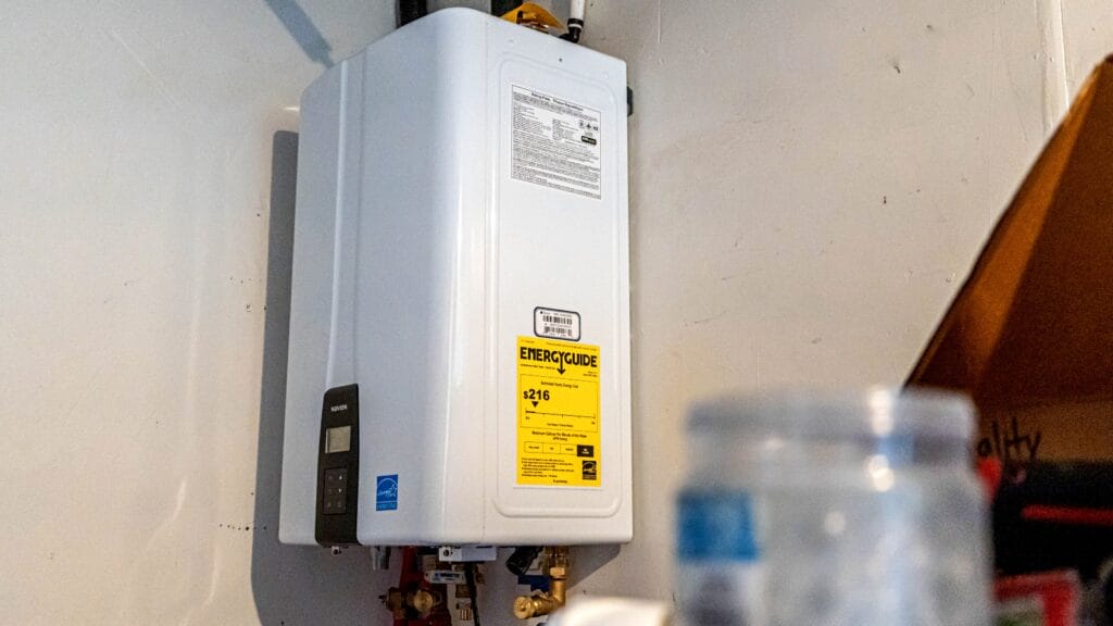 A close-up of a tankless water heater with an energy efficiency label. The surrounding space shows part of a utility room with plumbing and heating connections.