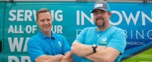 Two smiling Intown Plumbing technicians standing confidently in front of a branded service van, wearing blue company uniforms and crossing their arms.