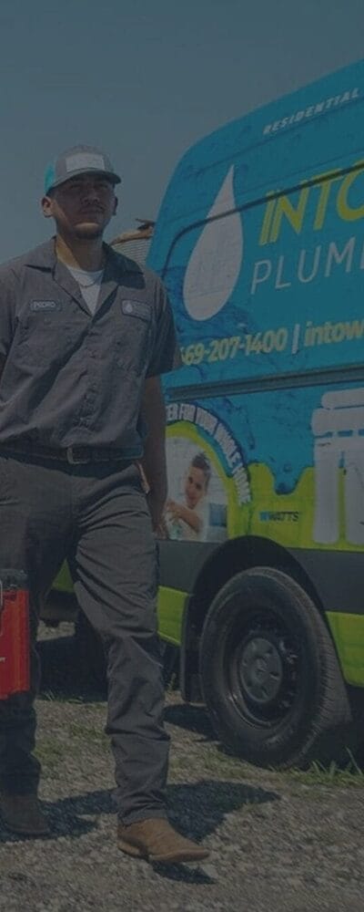 A professional plumber wearing a grey uniform stands next to a branded plumbing service van outdoors.