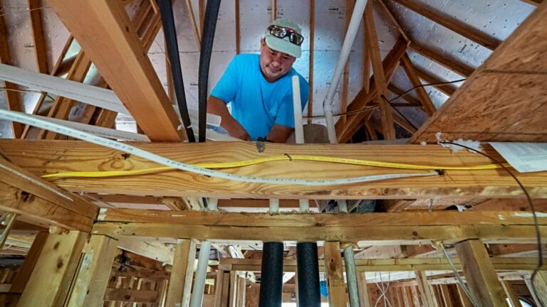 A plumber in a blue shirt works on pipes and electrical wiring within the wooden framing of a new construction project.