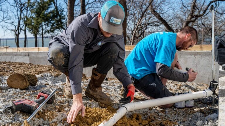 Two plumbers in work uniforms install white PVC piping at a new construction site with a concrete foundation.
