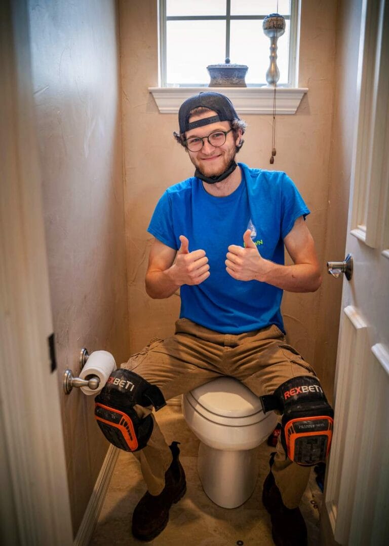 A smiling Intown Plumbing technician wearing knee pads and a blue uniform gives a thumbs up while sitting on a toilet during a repair job.