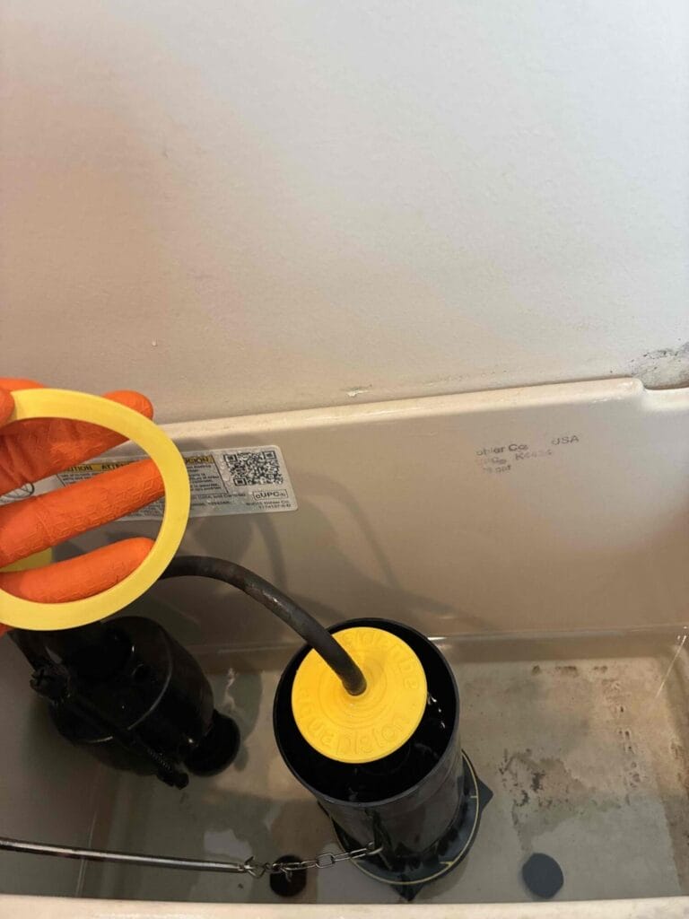 A plumber wearing orange gloves holds a new yellow toilet flapper seal while performing a toilet repair inside a tank.