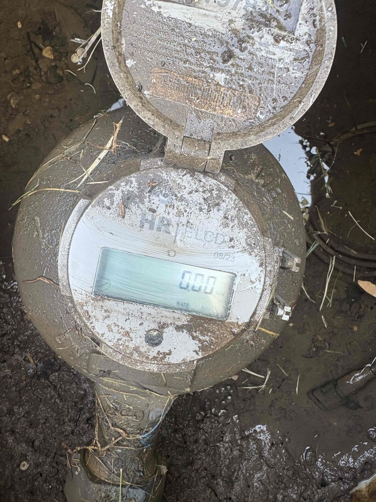 A close-up view of a digital water meter in a muddy ground box, showing a reading of 0.00 to help identify potential water leaks.