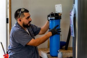 An Intown Plumbing technician wearing black gloves installs a blue commercial water filtration system in a workspace.