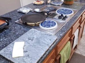 A kitchen island featuring a dark granite countertop, a built-in stove with decorative burner covers, a frying pan, and a white marble cutting board with a paper towel.