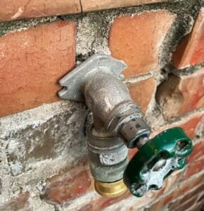 A close-up view of a metal outdoor water spigot mounted on a red brick wall, showing the valve handle and hose connection.