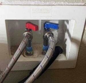 A white recessed washing machine water supply box featuring red and blue lever handles connected to braided metal hoses.