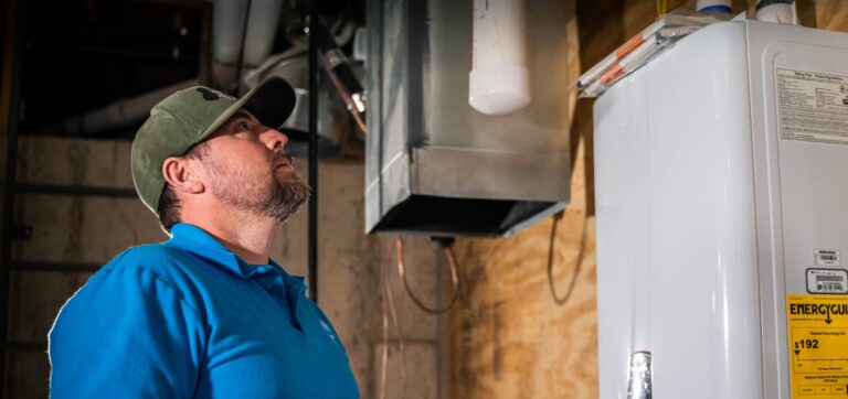 A professional plumber from Intown Plumbing wearing a blue polo shirt and green cap looks up at a wall-mounted tankless water heater while holding a smartphone to check for issues.