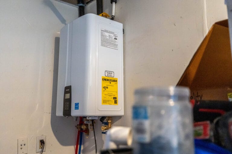 A white Navien tankless water heater mounted on a white wall with plumbing connections and an EnergyGuide yellow label visible.