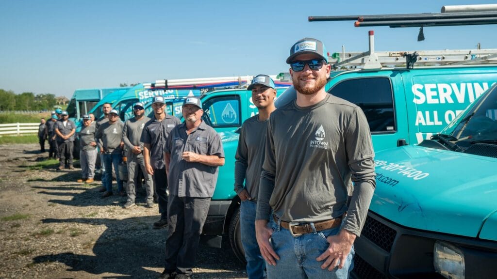 A group of Intown Plumbing technicians wearing branded shirts and hats standing in front of several company service vans.