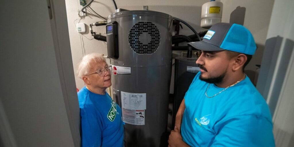 An elderly woman stands next to a young plumber and a large gray water heater in her home while he runs a water test.