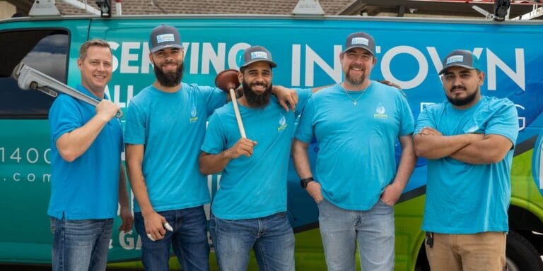 Five professional plumbers from Intown Plumbing standing in front of their company van, with one holding a pipe wrench and another holding a plunger.