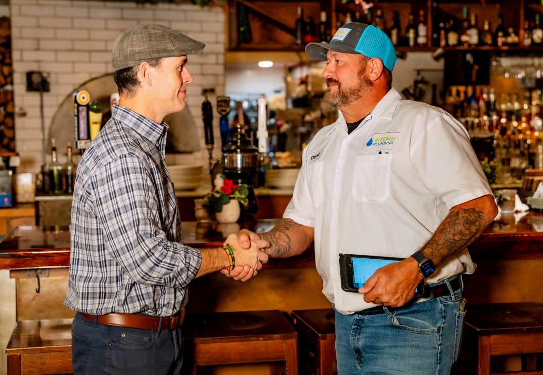 A professional plumber from Intown Plumbing wearing a branded shirt and cap shakes hands with a client inside a commercial establishment.