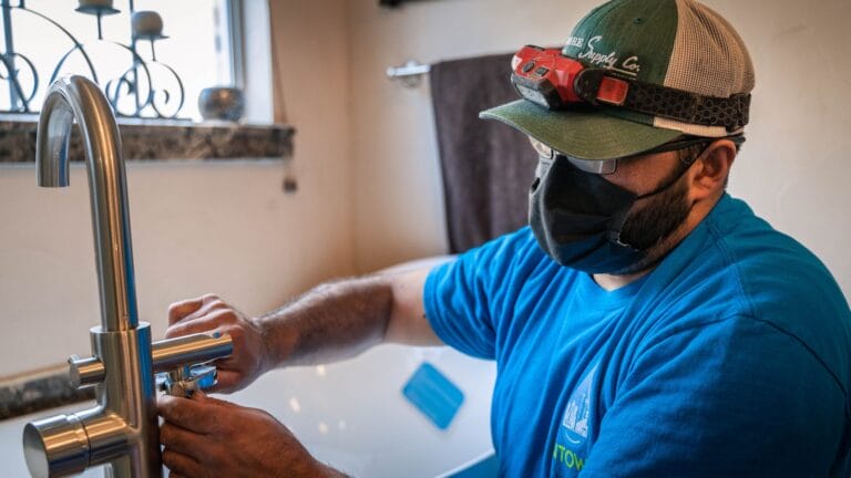 A professional plumber from Intown Plumbing wearing a headlamp and face mask installs a modern faucet in a bathroom.